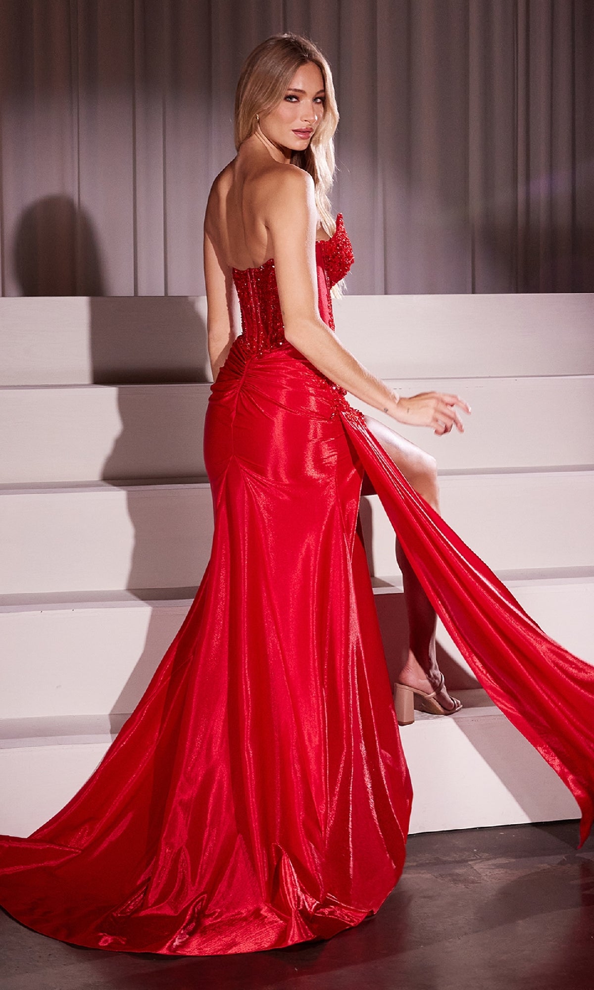 A woman in the Strapless Long Corset Prom Dress: Ladivine CM369 with a high slit stands on white steps, glancing back over her shoulder.